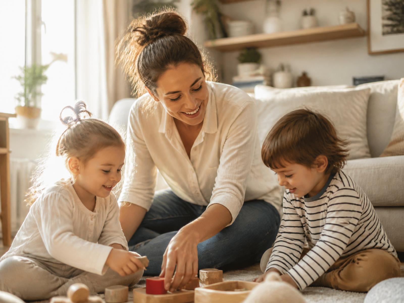 A babysitter playing on the floor with two happy young children in a sunlit living room